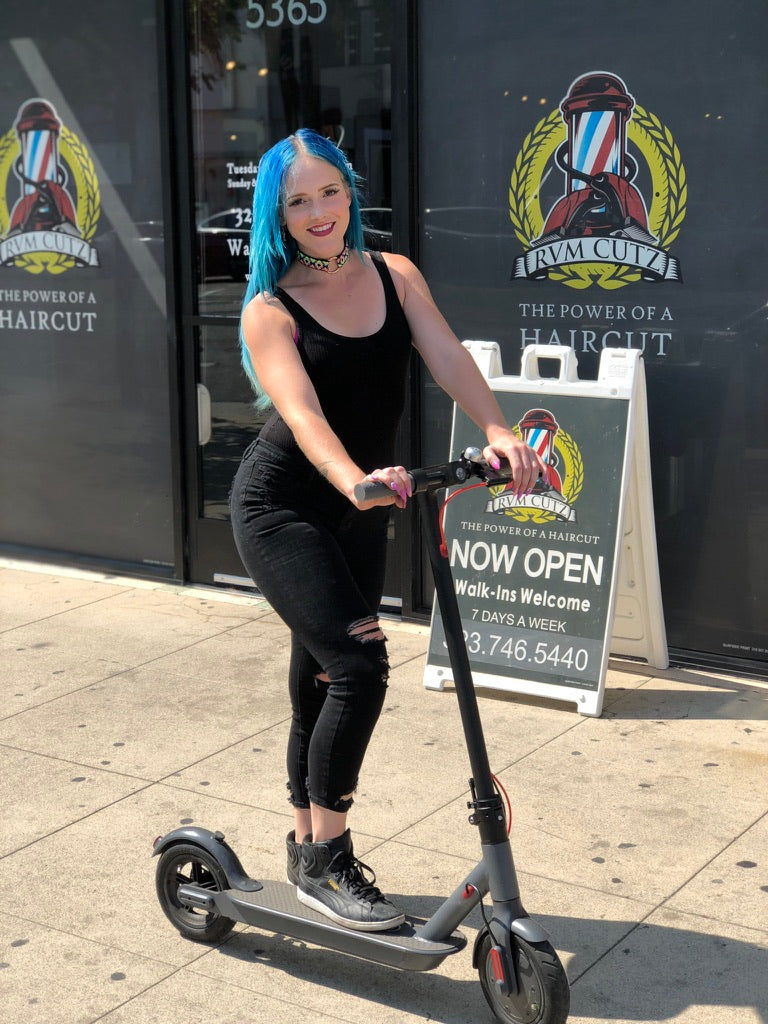 A blue-haired rider smiling on a Simply Brilliant scooter outside a storefront